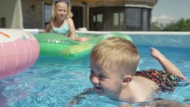 Lachend jongetje drijvend in het zwembad in de achtertuin om te leren zwemmen. Vrolijke peuter bij zijn eerste zwempartij in het zwembad. Verfrissende zomeractiviteiten met het gezin op warme zomerdagen. - Video
