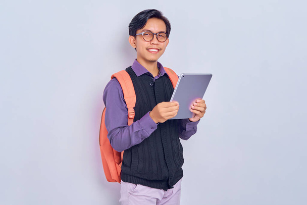 Gri arka planda izole edilmiş dijital tablet taşıyan sırt çantasıyla üniversiteden neşeli, genç Asyalı bir erkek öğrenci. Lise üniversite konsepti eğitimi - Fotoğraf, Görsel