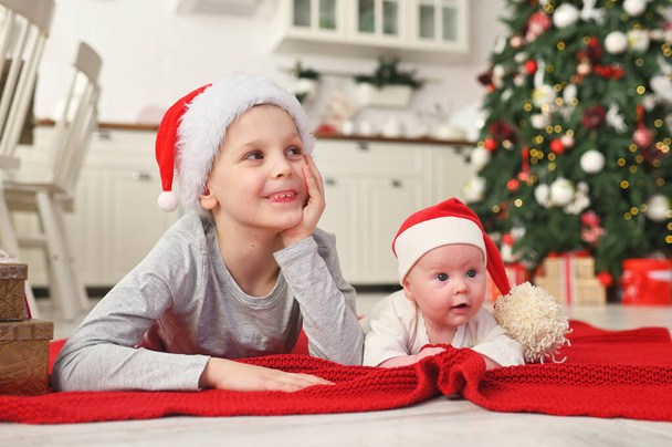 brother with little sister baby in Santa hats lie on a red blanket and smile, on the background of the Christmas tree. - Foto, imagen