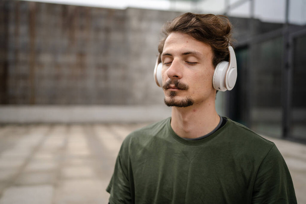 One man young adult caucasian male eyes closed for guided training yoga or meditation while sitting outdoor with headphones self-care practice real people well-being inner peace and balance concept - Photo, Image
