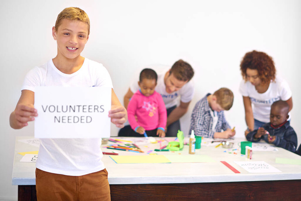 Wir brauchen immer mehr helfende Hände. Porträt eines Freiwilligen, der ein von Freiwilligen benötigtes Schild hält, mit Freiwilligen, die mit kleinen Kindern im Hintergrund arbeiten - Foto, Bild