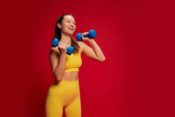 Portrait of young woman in yellow sportswear doing exercises with dumbbells isolated over red background. Strong hands. Concept of youth culture, emotions, facial expression, sport, lifestyle, health - Photo, Image