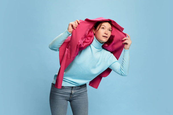 Portret van een jonge vrouw die poseert, hoofd bedekkend met roze jas geïsoleerd over blauwe achtergrond. Concept jeugdcultuur, emoties, gezichtsuitdrukking, mode, beroep, levensstijl - Foto, afbeelding