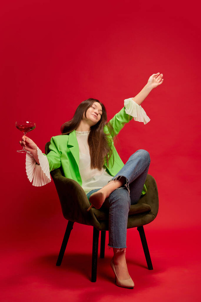 One young woman posing in stylish clothes, sitting on chair with cocktail and enjoying weekends isolated over red background. Concept of youth culture, emotions, facial expression, fashion, party - Photo, image