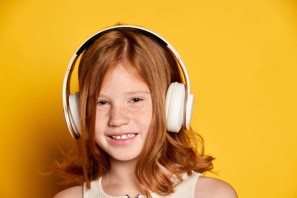 Open eyesight. Closeup portrait of little stylish girl with freckles wearing headphones isolated over bright yellow background. Concept of children emotions, dance, music, happiness. Copy space for ad - 写真・画像