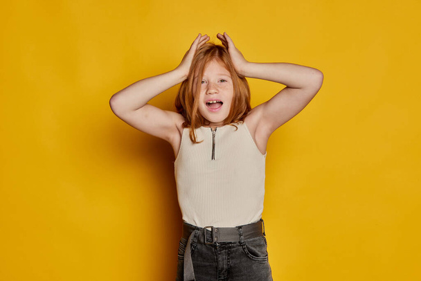 Wow, shock, surprise. Little stylish school age girl with long red hair and freckles isolated over bright yellow background. Concept of children emotions, dance, music, happiness. Copy space for ad - Foto, Imagen