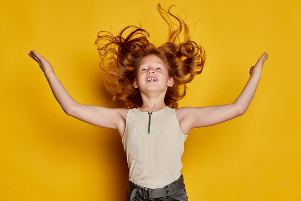 Dance and delight. Excited little stylish girl with long red hair and freckles isolated over bright yellow background. Concept of children emotions, dance, music, happiness. Copy space for ad - Foto, Bild