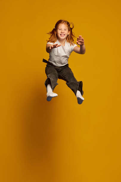 Flight. Astonished little girl, kid in white t-shirt and jeans jumping high isolated over yellow background. Kids fashion, emotions, carefree childhood, challenges, education concept - Photo, image