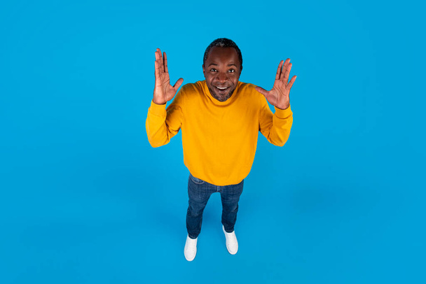 High angle view of emotional excited handsome middle aged black man in stylish casual outfit raising hands up and smiling isolated on blue studio background, full length shot, copy space - Fotoğraf, Görsel