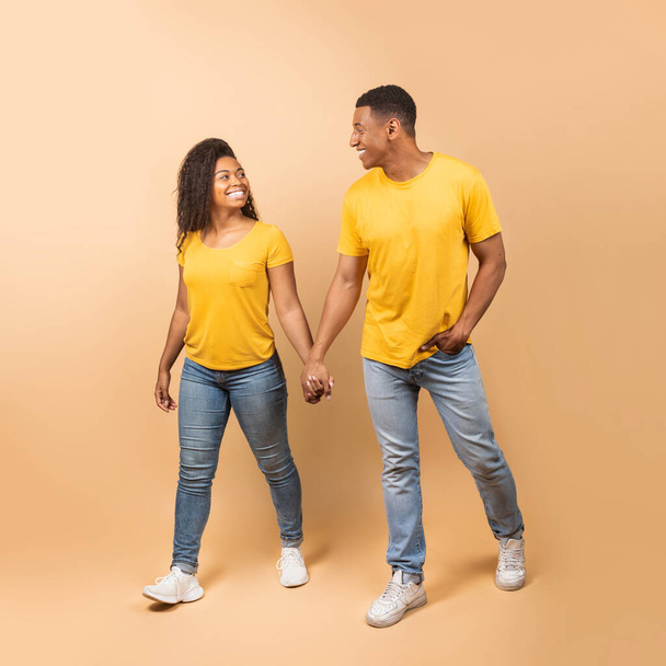 Romantic african american couple walking, holding hands, looking at each other and smiling on peach studio background, full length. Young black boyfriend and girlfriend going for walk together - Photo, image