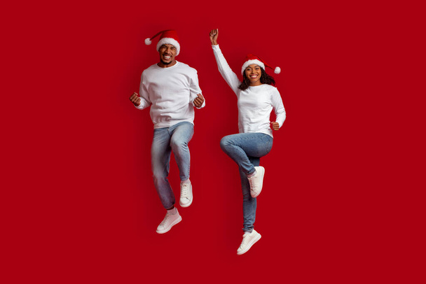 Happy african american xmas lovers millennial man and woman in casual having fun on red studio background, wearing Santa hats, jumping in the air, gesturing and smiling, copy space, full length - Fotoğraf, Görsel