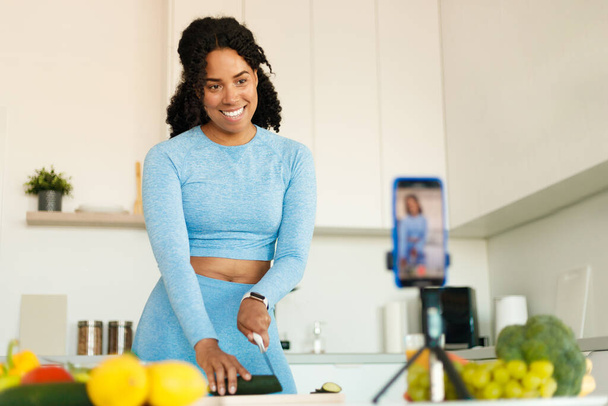 Femme noire excitée en tenue de sport enregistrement vidéo sur caméra smartphone, préparation de salade fraîche dans la cuisine, couper des légumes et parler à la caméra du téléphone - Photo, image