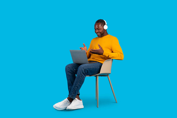 Cheerful mature black man in stylish casual outfit sitting on chair isolated on blue studio background, using modern laptop and wireless stereo headphones, have video call, copy space, full length - Photo, Image