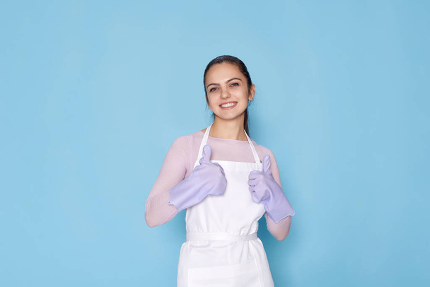 beautiful smiling woman in purple rubber gloves and cleaner apron showing ok gesture on blue background. cleaning - Fotografie, Obrázek