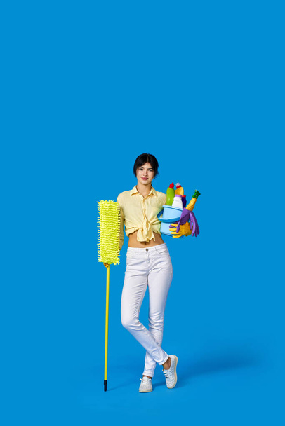 koncentrovaná vážná hospodyňka v gumových rukavicích, úklid s mopem a držící vědro detergentů na modrém pozadí. Plná délka - Fotografie, Obrázek