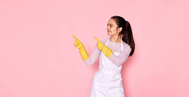 beautiful caucasian woman in rubber gloves pointing fingers aside at copy space on pink background. cleaning - Foto, Imagen