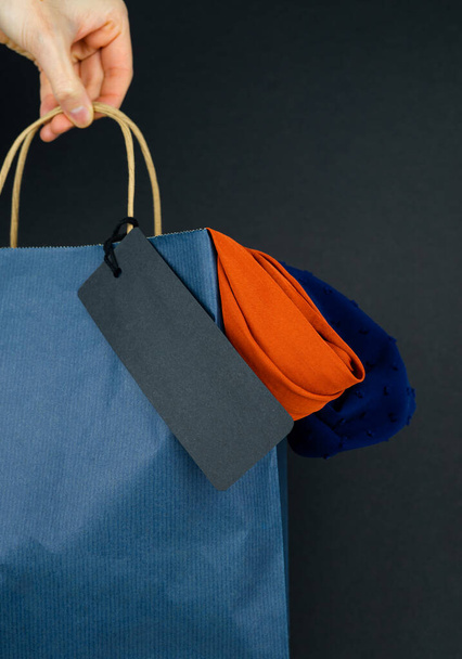 濃い背景に文字のない黒ラベルの青い紙のバッグを持つ女性の手。販売の概念。コピースペース。新年の売り上げ。垂直写真. - 写真・画像