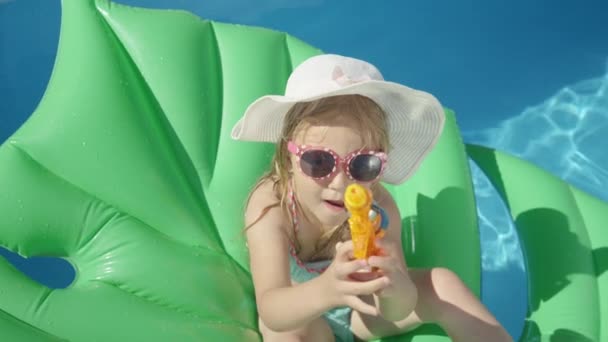 CLOSE UP Little girl sitting on a floatie with water gun pointing towards camera. Cheerful little girl floating in backyard swimming pool and having fun with water blaster spraying in camera direction - Footage, Video