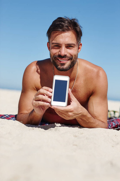 Este es el lugar perfecto. un joven mostrando una pantalla de móvil en blanco mientras está en la playa - Foto, imagen