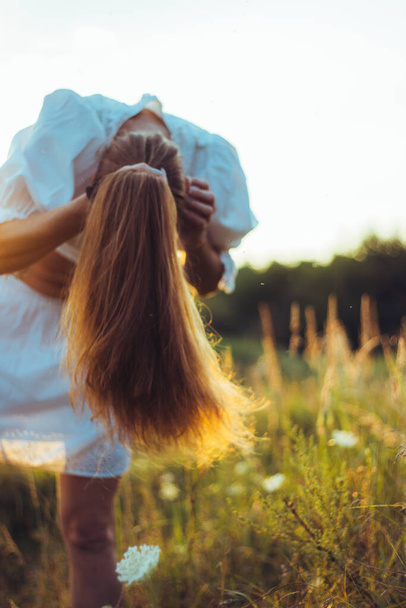 beautiful long-haired woman bent over to tie her hair in a sunny summer field. The concept of luxurious long red healthy hair, natural care and nurturing. Natural sunset light, selective focus - Фото, изображение