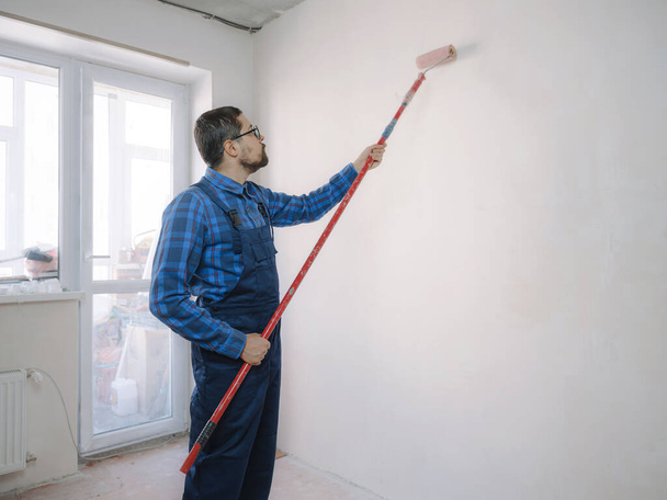 fiatal férfi kék munkaruhában tartja festék görgő, felett fehér fal szoba háttér. Műszerek tartozékok felújítás apartman szoba. A javítóotthon koncepciója - Fotó, kép