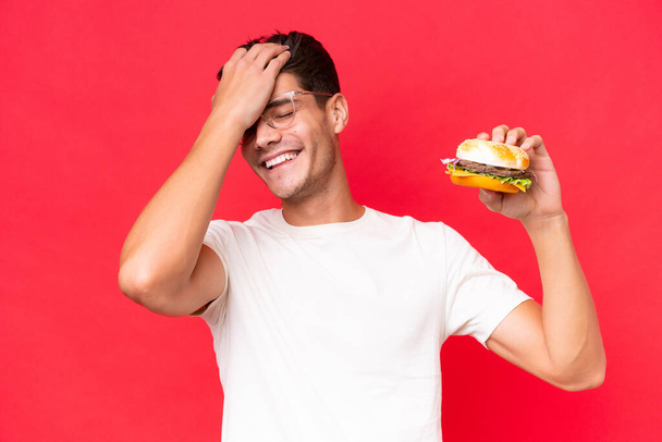 Mladý běloch, který drží burger izolovaný na červeném pozadí, si něco uvědomil a zamýšlí řešení - Fotografie, Obrázek