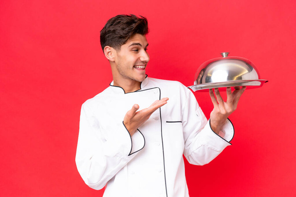 Joven chef caucásico hombre sosteniendo bandeja aislada sobre fondo rojo extendiendo las manos a un lado para invitar a venir - Foto, imagen