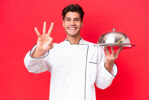 Молодий кавказький шеф-кухар тримає лоток ізольований на червоному тлі щасливий і рахуючи трьох пальцями
 - Фото, зображення