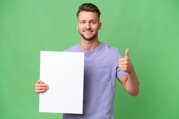 Young blonde caucasian man over isolated background holding an empty placard with thumb up - Zdjęcie, obraz