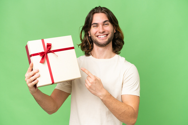 Genç yakışıklı bir adam, izole bir arka plan üzerinde bir hediye tutuyor ve onu işaret ediyor. - Fotoğraf, Görsel