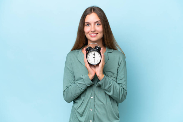 Młoda piękna kobieta na niebieskim tle w piżamie i trzymając zegar ze szczęśliwym wyrazem - Zdjęcie, obraz