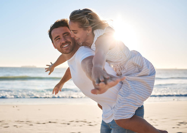 Happy, couple and piggyback walk on the beach for love, travel or summer vacation bonding together in the outdoors. Man carrying woman on back with smile enjoying playful fun time walking by the sea. - Photo, Image