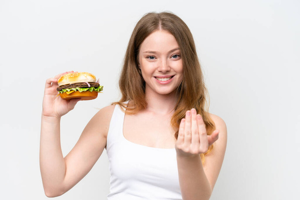 Jonge mooie vrouw met een hamburger geïsoleerd op witte achtergrond die uitnodigt om met de hand te komen. Blij dat je gekomen bent. - Foto, afbeelding