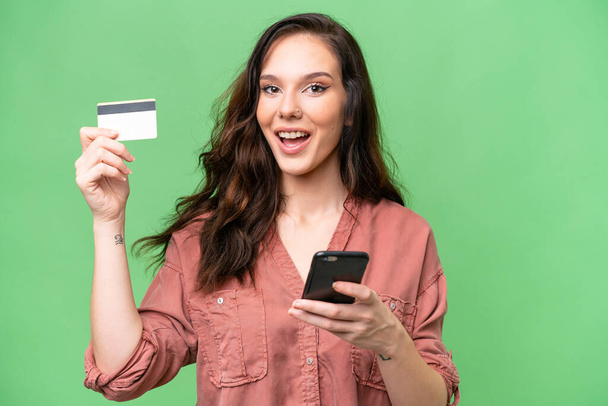 Joven mujer caucásica sobre la compra de fondo aislado con el móvil y la celebración de una tarjeta de crédito con expresión sorprendida - Foto, Imagen