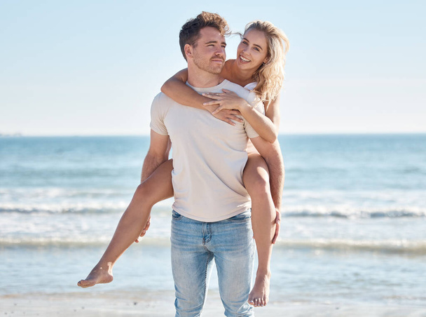Αγάπη, καλοκαίρι και ζευγάρι κάνουν piggyback στην παραλία απολαμβάνοντας ρομαντικές διακοπές, διακοπές και μήνα του μέλιτος από τη θάλασσα. Φύση, στοργή και άντρας που κουβαλάει γυναίκα δίπλα στον ωκεανό, που δένεται χαμογελώντας και ευτυχισμένα μαζί. - Φωτογραφία, εικόνα