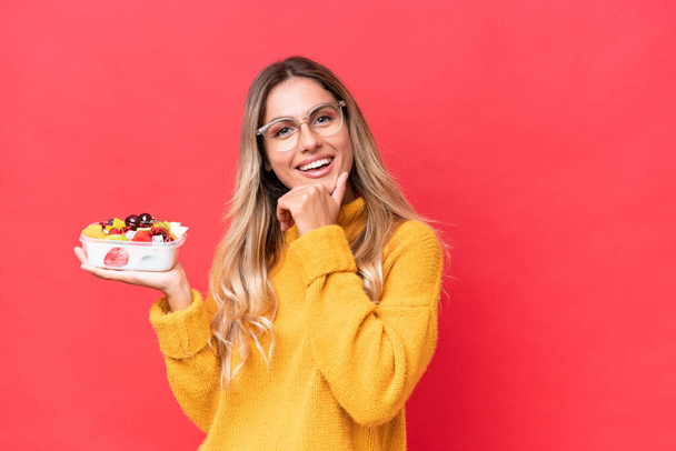 Genç, güzel Uruguaylı bir kadın elinde bir kase meyveyle kırmızı arka planda mutlu ve gülümsüyor. - Fotoğraf, Görsel