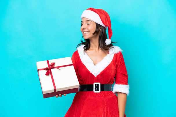 Young hispanic woman dressed as mama noel holding a gift isolated on blue background looking to the side and smiling - Photo, Image