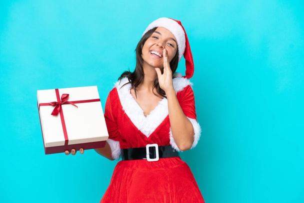 Anne Noel gibi giyinmiş genç İspanyol kadın elinde mavi arka planda izole edilmiş bir hediyeyle ağzı açık bağırıyor. - Fotoğraf, Görsel