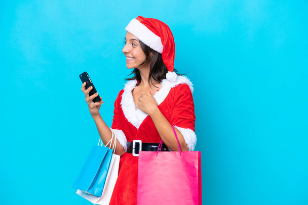 Anne Noel gibi giyinmiş genç İspanyol kadın mavi arka planda izole edilmiş. Elinde alışveriş çantaları ve cep telefonu var. - Fotoğraf, Görsel