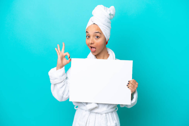 Młoda kobieta w szlafroku z ręcznikiem odizolowanym na niebieskim tle trzymająca pusty plakat i robiąca znak OK - Zdjęcie, obraz