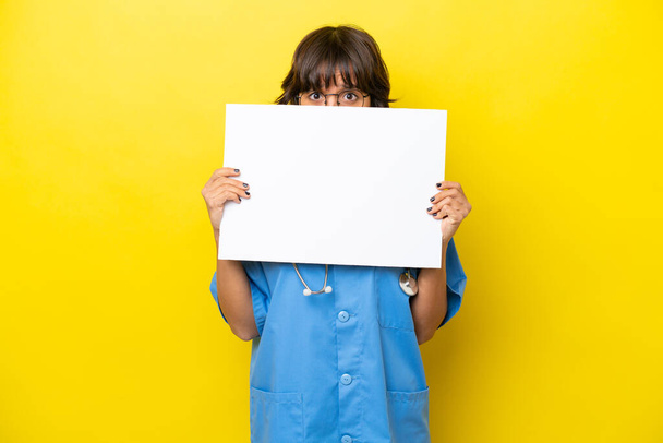 Young nurse doctor woman isolated on yellow background holding an empty placard and hiding behind it - Photo, Image