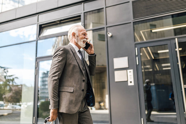 Hymyilevä liike-elämän ammattilainen puhuu älypuhelimella. Iäkkäiden miesyrittäjien sivunäkymä kulkee kaupungin modernin toimistorakennuksen ohi. Hänellä on täysi puku.. - Valokuva, kuva