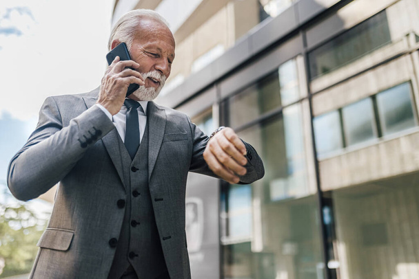 Senior-Geschäftsmann überprüft die Uhrzeit auf der Armbanduhr. Der männliche Profi im Anzug telefoniert mit dem Smartphone. Er stellt sich gegen Firmengebäude. - Foto, Bild