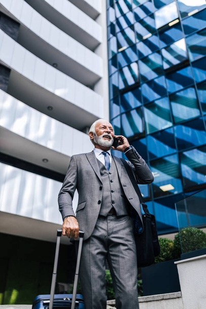 Low angle view of smiling senior professional talking on smart phone. Businessman wearing suit is carrying laptop bag and holding suitcase. He is standing against office building in the city. - Foto, Bild