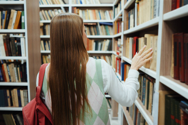 Female student walking among rows of bookcases with backpack on shoulder, touching some of book covers. Woman in library. Concept of education - Photo, image