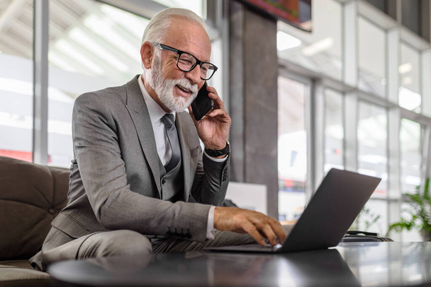 Selbstbewusster Unternehmer im Smartphone-Gespräch. Lächelnde männliche Seniorprofis arbeiten am Laptop am Tisch. Er sitzt im vollen Anzug im Wartezimmer des Bahnhofs. - Foto, Bild
