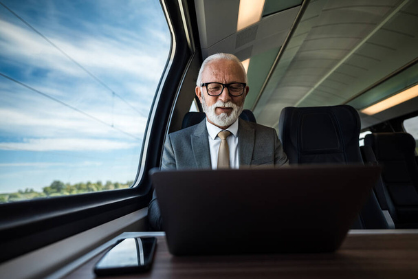 Lächelnder Geschäftsmann, der am Laptop arbeitet, während er im Personenzug am Fenster sitzt. Der ältere Unternehmer trägt Brille und Anzug. Auf Geschäftsreisen ist er zuversichtlich. - Foto, Bild
