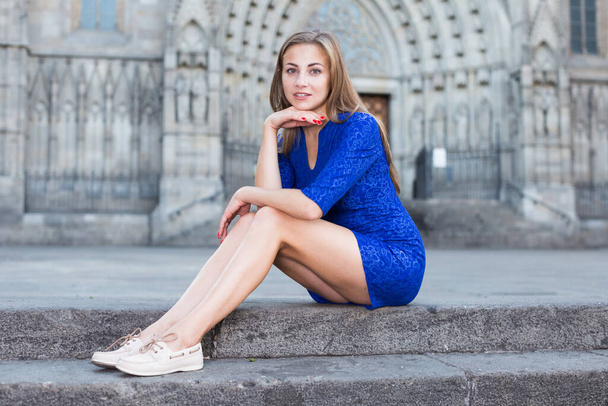 Cheerful female is playfully posing sitting in blue dress on the stairs outdoor. - Photo, Image