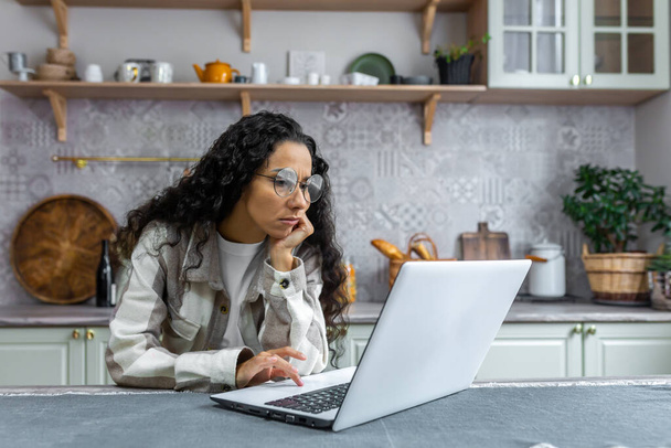 Psychologist online. Young hispanic woman sitting sad at home in the kitchen in a depressed state. Consults with a psychologist online from a laptop. - Foto, Bild