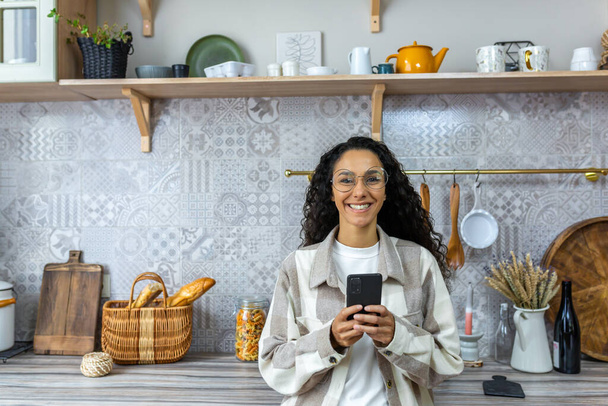 A beautiful young Latin American woman is holding a phone in her hands. Standing at home in the kitchen, looking at the camera, smiling. - Photo, image